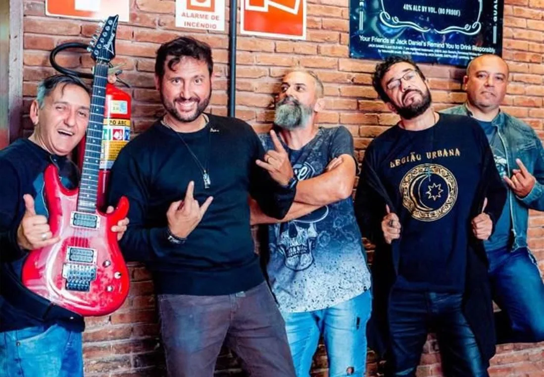 Integrantes da banda Legião Urbana Cover posam descontraídos em frente a uma parede de tijolos, com gestos rock'n'roll e expressão irreverente. Um dos músicos segura uma guitarra vermelha, enquanto outro veste uma camiseta com o nome e símbolo da banda homenageada.