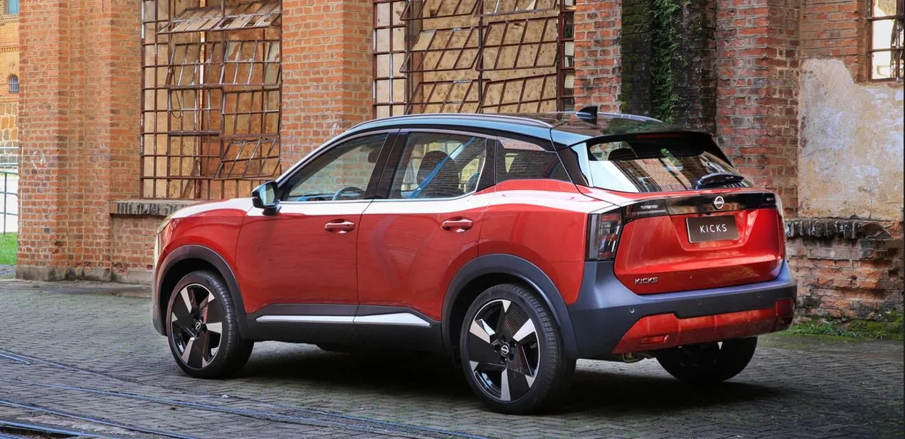 Novo Nissan Kicks 2025 visto de traseira em ambiente urbano com arquitetura de tijolos à mostra, destacando a nova lanterna em LED conectada por faixa escurecida, acabamento biton com carroceria vermelha e teto preto, além das rodas aro 19" com design exclusivo, evidenciando o estilo moderno e arrojado do SUV compacto nacional.
