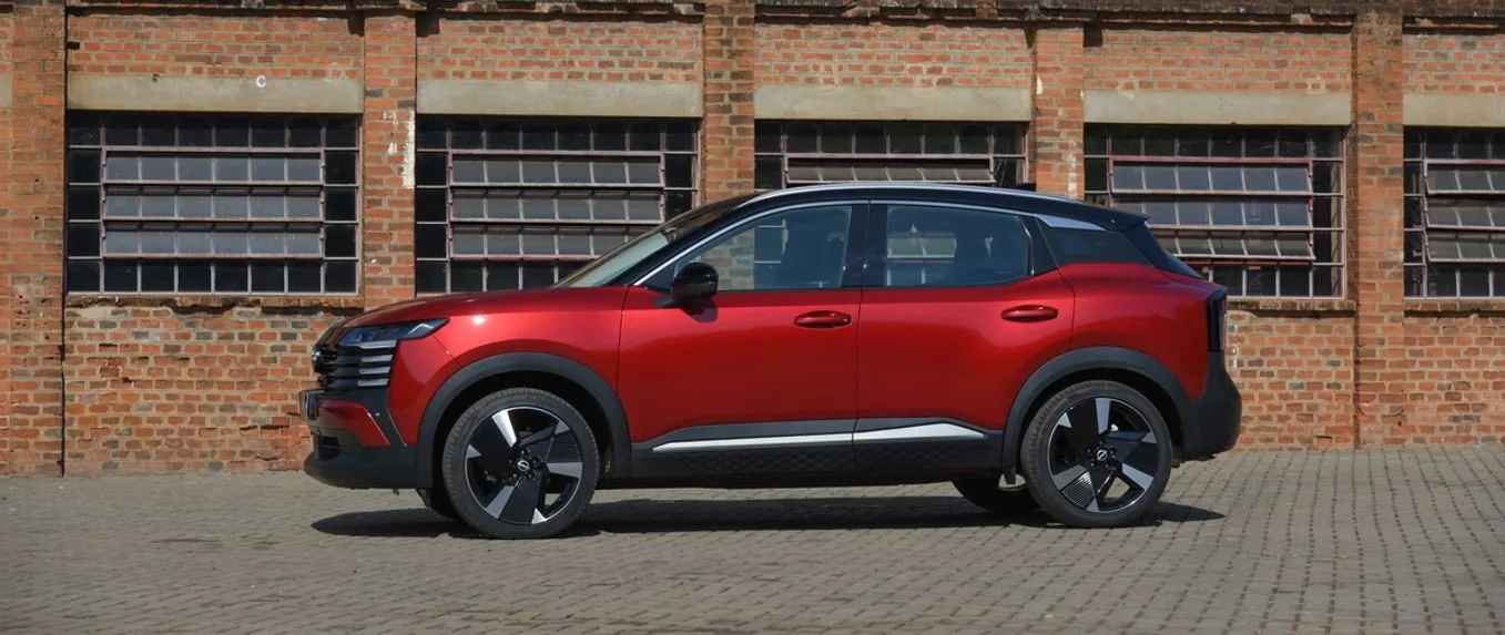 Vista lateral do novo Nissan Kicks 2025 estacionado em frente a uma parede de tijolos, destacando o design renovado com linhas fluidas, pintura biton em vermelho e teto preto, rodas de liga leve aro 19" com acabamento diamantado e molduras de caixa de roda em preto fosco, evidenciando a proposta urbana, moderna e robusta do SUV compacto nacional.