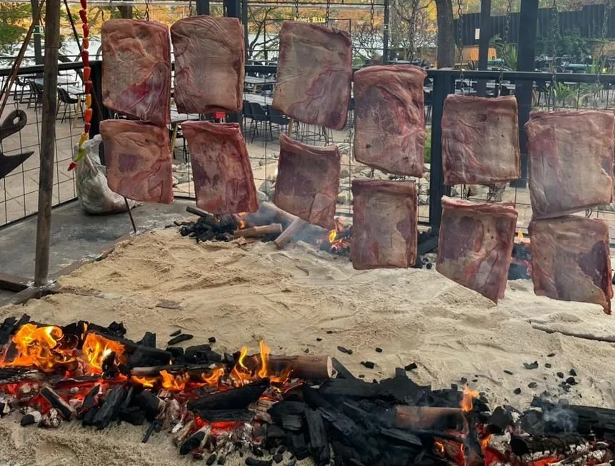 Costelas bovinas cruas penduradas em estrutura de ferro sendo assadas lentamente em fogo de chão.