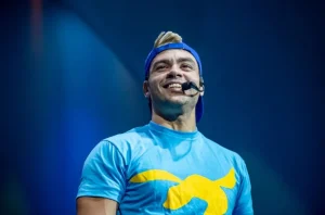 Luccas Neto sorri durante apresentação do espetáculo “Luccas e Gi em: O Mundo de Magia e Fantasia” em Belo Horizonte, usando microfone auricular e camiseta azul com símbolo amarelo, sob iluminação cênica.