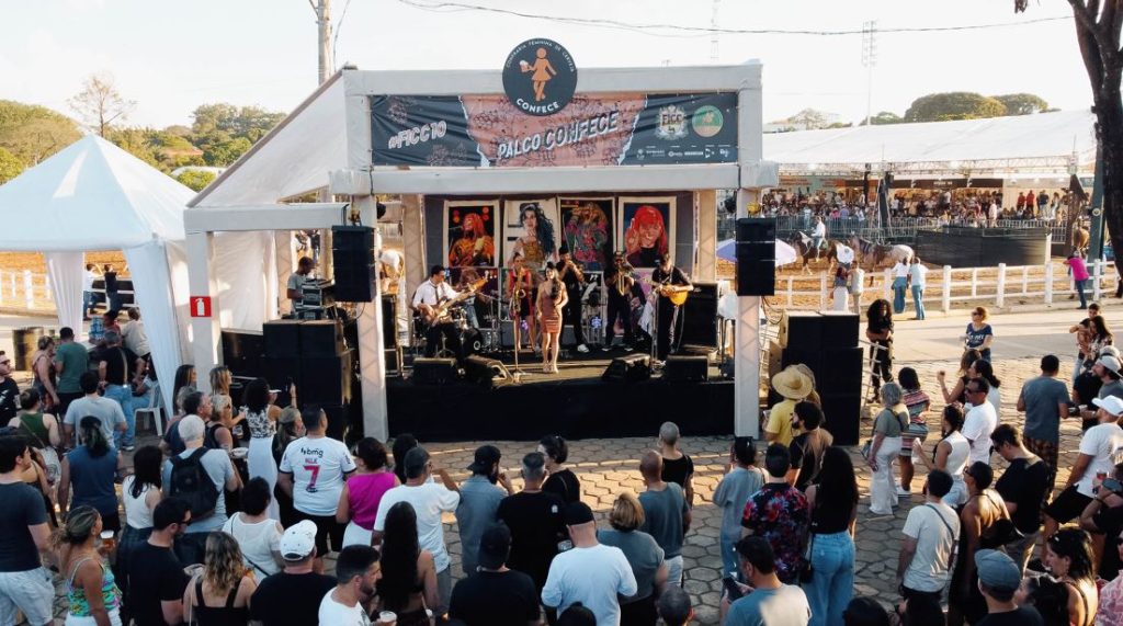 Plateia acompanha apresentação ao vivo no Palco Confece durante o FICC, com banda performando em frente a painéis artísticos coloridos.