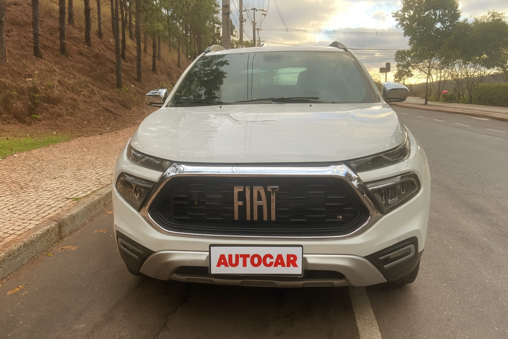 Vista frontal da Fiat Toro Ranch 2.2 Turbodiesel branca, estacionada ao lado de uma calçada arborizada. A imagem destaca a grade dianteira com moldura cromada e o logotipo central da Fiat em bronze, além dos faróis full LED com desenho arrojado.