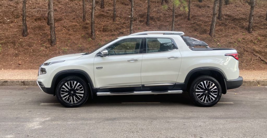 Vista lateral completa da Fiat Toro Ranch 2.2 Turbodiesel na cor branca, estacionada em via urbana com fundo arborizado. A imagem destaca o perfil elegante da picape com rodas de 18 polegadas, estribos cromados, santantônio integrado e linhas de carroceria bem definidas.