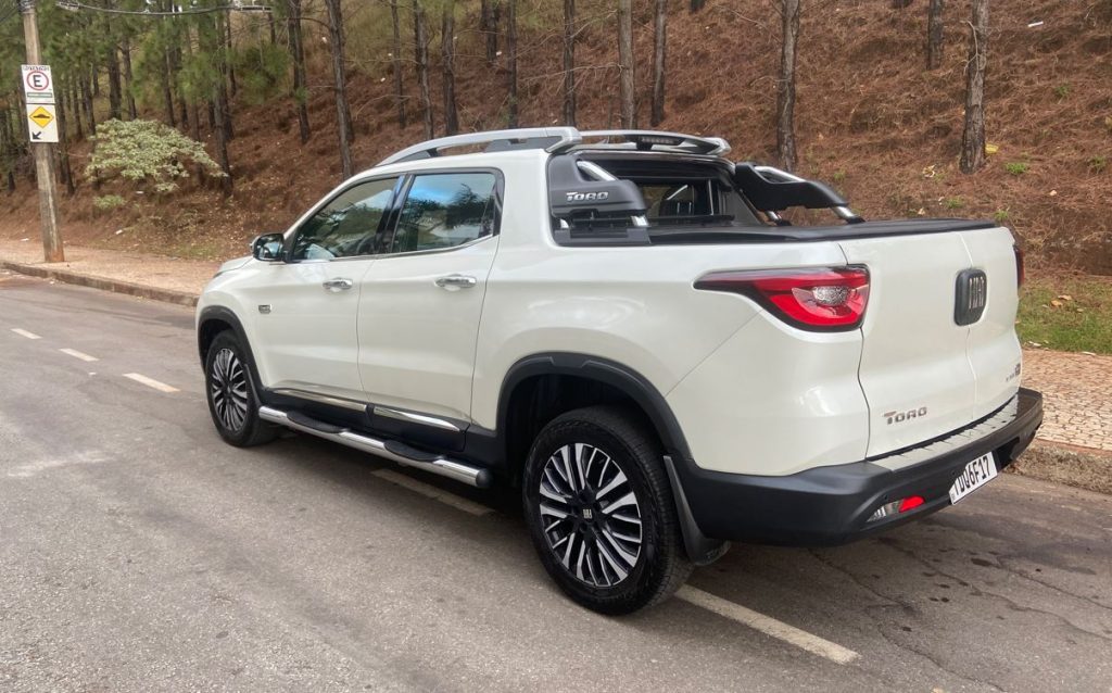 Vista traseira lateral da Fiat Toro Ranch 2.2 Turbodiesel na cor branca, estacionada em rua arborizada. A imagem destaca as lanternas em LED, o santantônio robusto com o nome 'Toro' em destaque, as rodas diamantadas de 18 polegadas e os estribos laterais cromados.