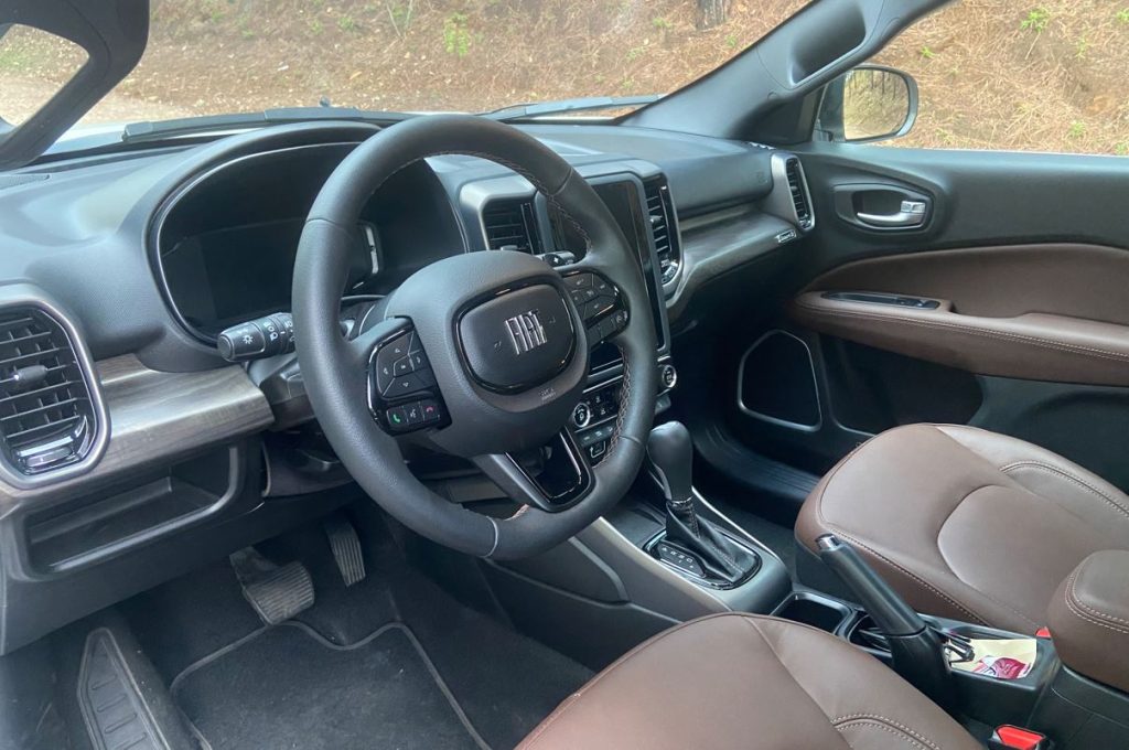 Interior da Fiat Toro Ranch 2.2 Turbodiesel com acabamento premium. Volante multifuncional com logo Fiat, bancos em couro marrom com costuras aparentes, central multimídia vertical e detalhes em tons escurecidos no painel e nas portas, transmitindo requinte e modernidade.