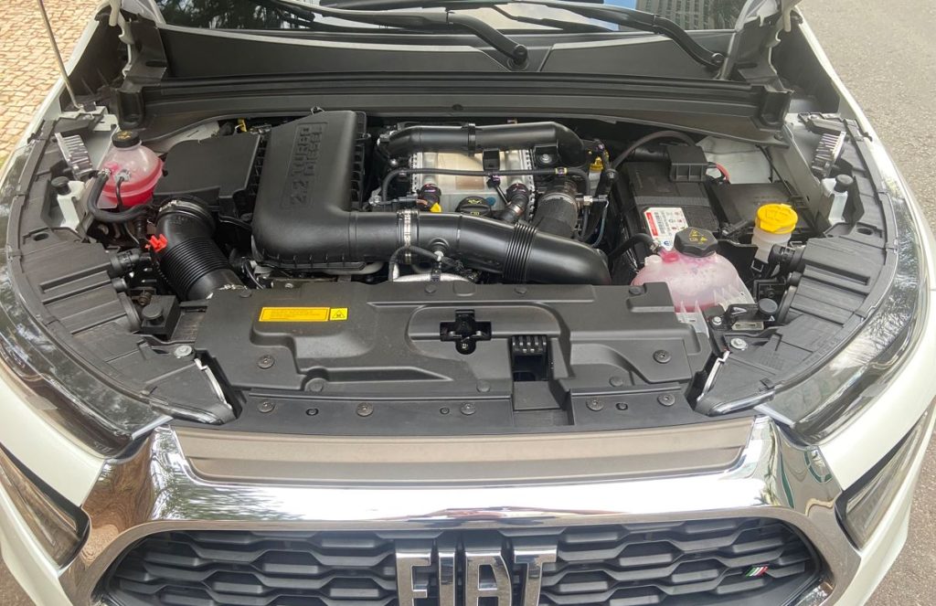 Imagem do compartimento do motor da Fiat Toro Ranch 2.2 Turbodiesel com o capô aberto. O motor Multijet 2.2 aparece em destaque, com componentes modernos como o turbo com geometria variável, mangueiras reforçadas, reservatórios identificados e estrutura bem organizada.