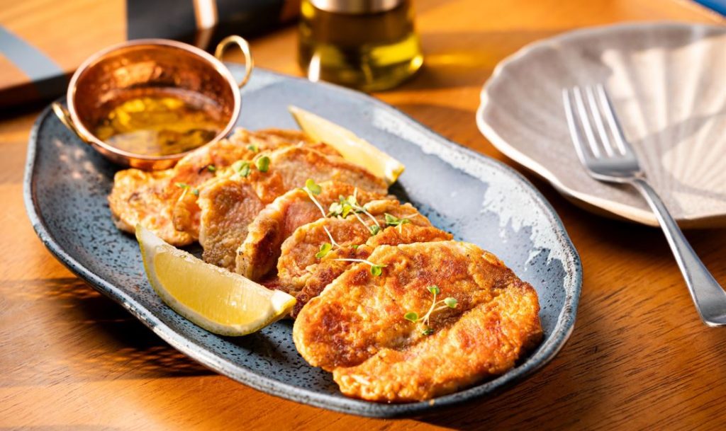 Filés de peixe grelhado na manteiga, servidos em prato cerâmico com limão siciliano, brotos verdes e molho à parte.