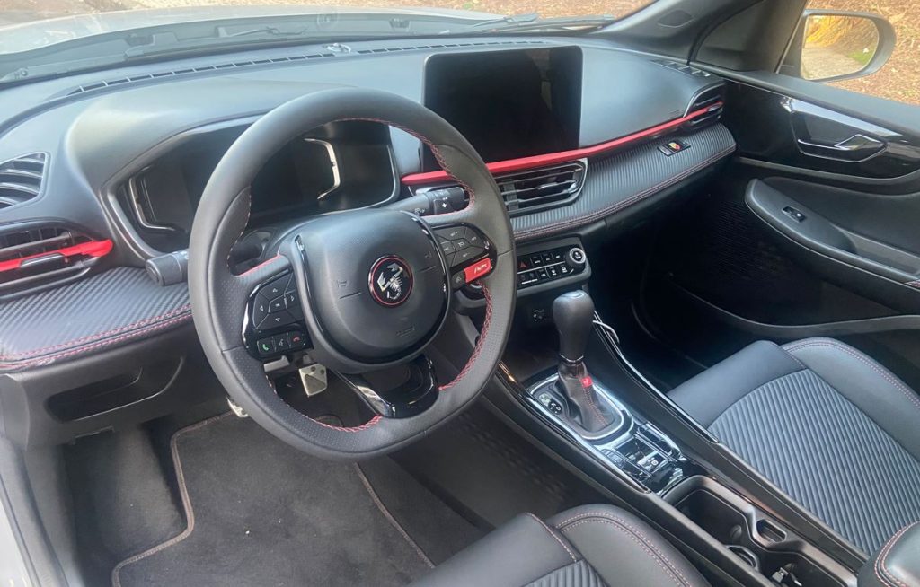 Interior do Fastback Abarth 2025 com volante esportivo multifuncional, costuras vermelhas e painel com acabamento em fibra de carbono.