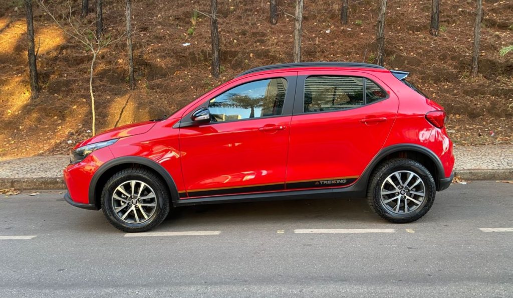 Vista lateral do Fiat Argo Trekking 2026 na cor vermelha, exibindo rodas de liga-leve, molduras nas caixas de roda e faixa lateral exclusiva "Trekking", estacionado em via urbana ladeada por árvores e calçada de pedras portuguesas.