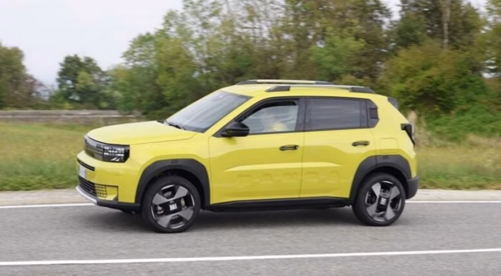 Fiat Grande Panda 2026 em movimento em uma estrada asfaltada, exibindo sua carroceria amarela vibrante e design robusto estilo mini-SUV, com vegetação ao fundo e céu nublado.