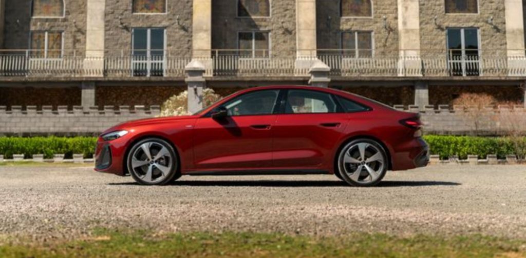 Vista lateral do novo Audi A5 2025 em vermelho, destacando o perfil fastback, rodas esportivas e design elegante diante de casarão clássico.