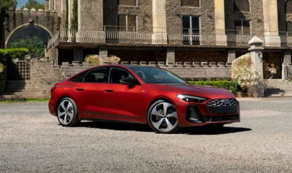 Novo Audi A5 2025 em cor vermelho granadina estacionado em frente a construção histórica, destacando linhas elegantes e rodas esportivas.