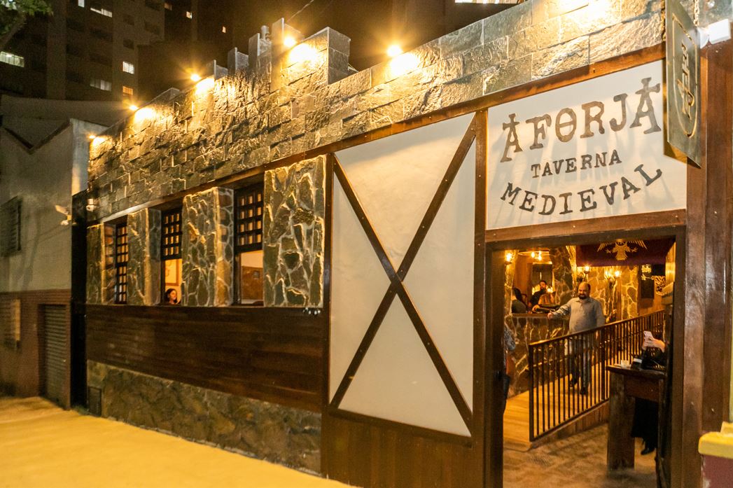 Fachada iluminada da A Forja Taverna Medieval em Belo Horizonte, com entrada em estilo rústico e detalhes que remetem à arquitetura medieval.
