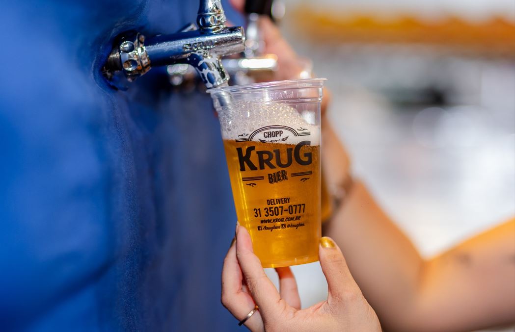 Pessoa serve chope claro da Krug Bier em copo plástico transparente, diretamente de uma torneira metálica durante a Oktoberfest BH.