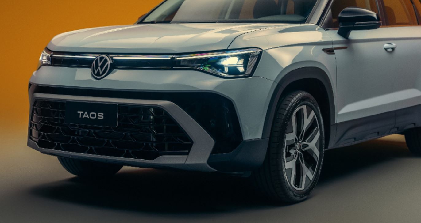 Detalhe da dianteira do Novo Volkswagen Taos 2026 com grade frontal redesenhada, faróis IQ.Light em LED e rodas de liga leve diamantadas, em fundo laranja de estúdio.