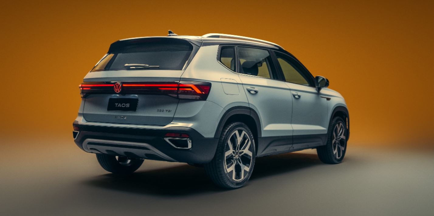 Traseira do Novo Volkswagen Taos 2026 com logo iluminado em LED vermelho, novas lanternas conectadas por faixa luminosa e acabamento esportivo em destaque, sob fundo laranja vibrante.