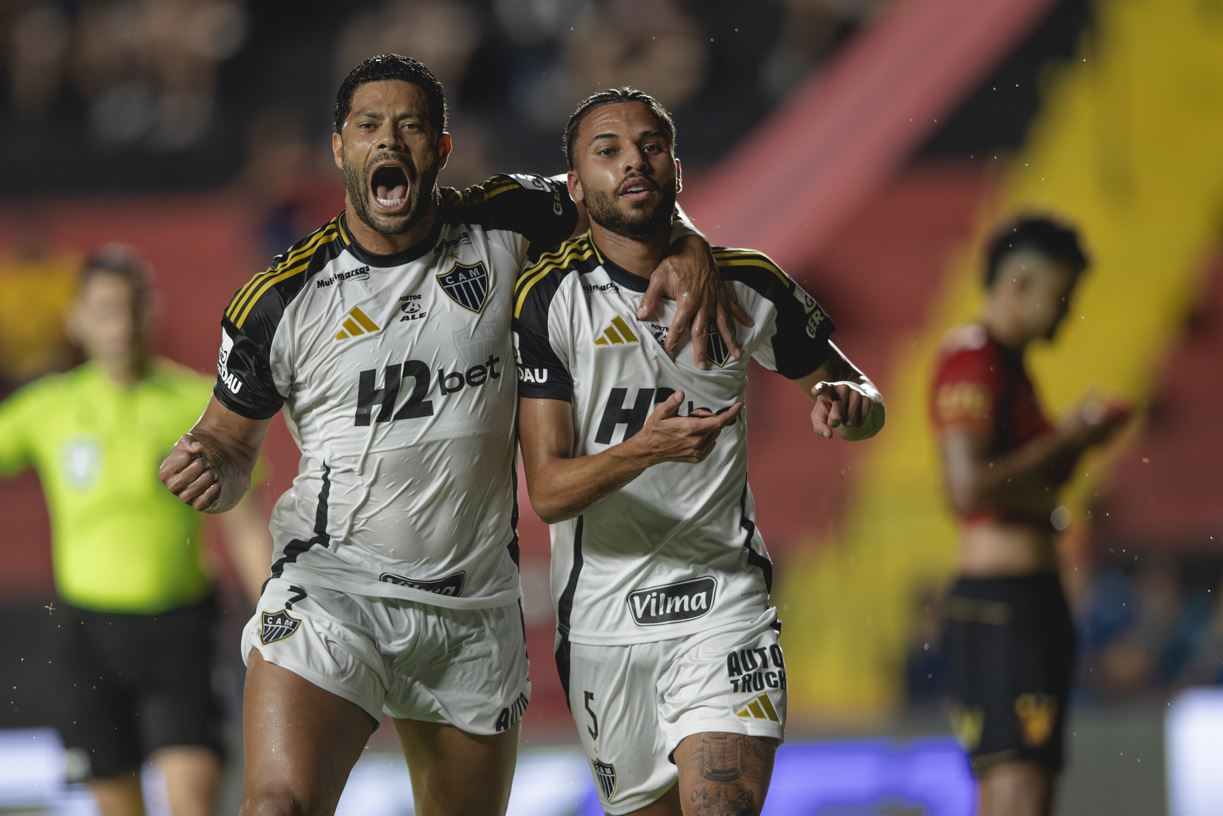 Hulk e Alexsander, jogadores do Atlético Mineiro, comemoram abraçados após gol marcado contra o Sport Recife, em partida válida pela 33ª rodada do Campeonato Brasileiro, com expressão de euforia e intensidade, enquanto jogador adversário aparece ao fundo, cabisbaixo.