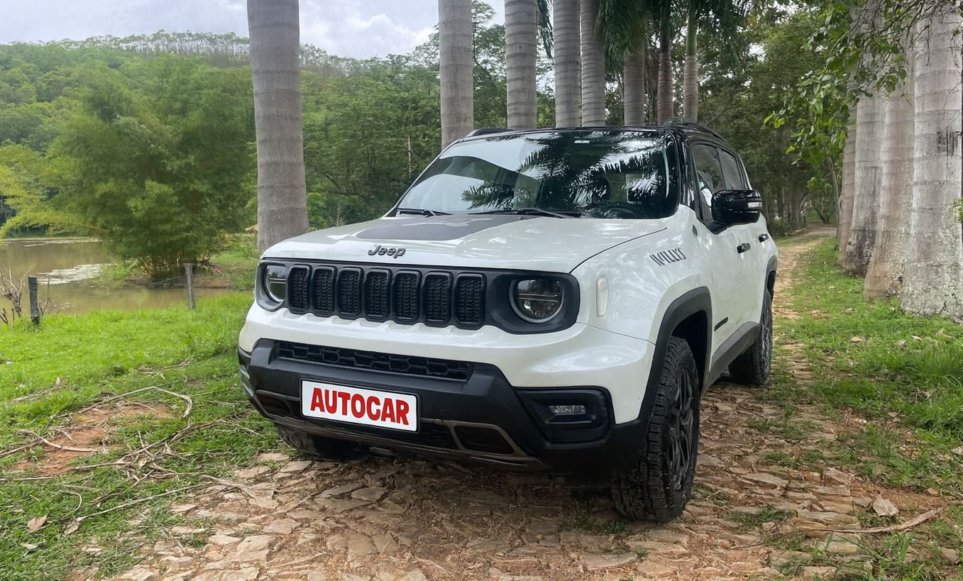Jeep Renegade Willys 10 Anos branco em estrada de terra cercada por palmeiras e vegetação, com lago ao fundo em área rural.