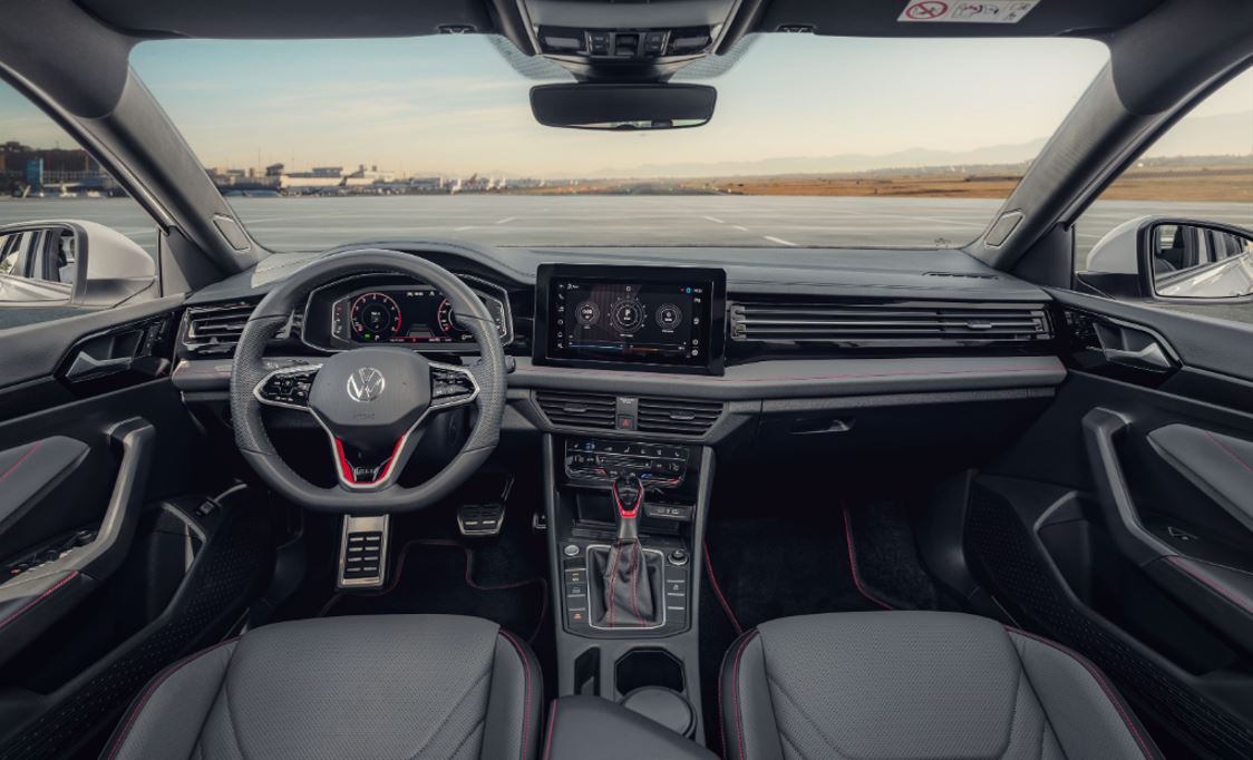 Painel do Volkswagen Jetta GLI 2026 visto do banco traseiro, com acabamento esportivo, volante multifuncional com base reta, costuras vermelhas contrastantes, quadro de instrumentos digital de 10,25", central multimídia VW Play de 10", comandos do ar-condicionado digital Climatronic Touch, alavanca de câmbio DSG e detalhes em preto brilhante.