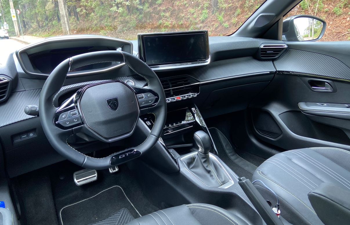 Interior do Peugeot 208 GT híbrido com volante esportivo em couro, central multimídia flutuante e acabamento estilo fibra de carbono.