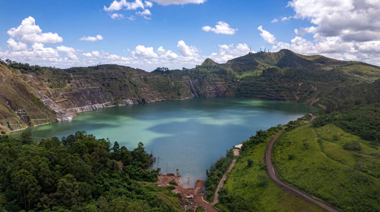 Vista panorâmica da cava da antiga Mina de Águas Claras, em Nova Lima, com grande lago de águas verdes cercado por encostas rochosas e vegetação nativa, sob céu azul com nuvens esparsas.