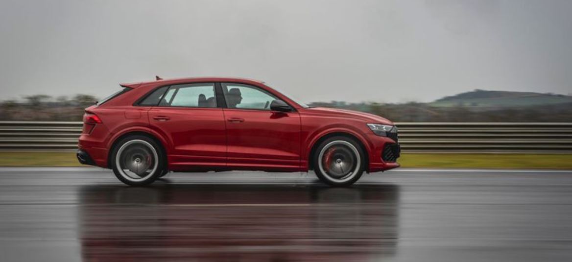 Audi RS Q8 Performance 2026 em vista lateral, na cor vermelha, em alta velocidade sobre pista molhada com fundo de paisagem nublada.