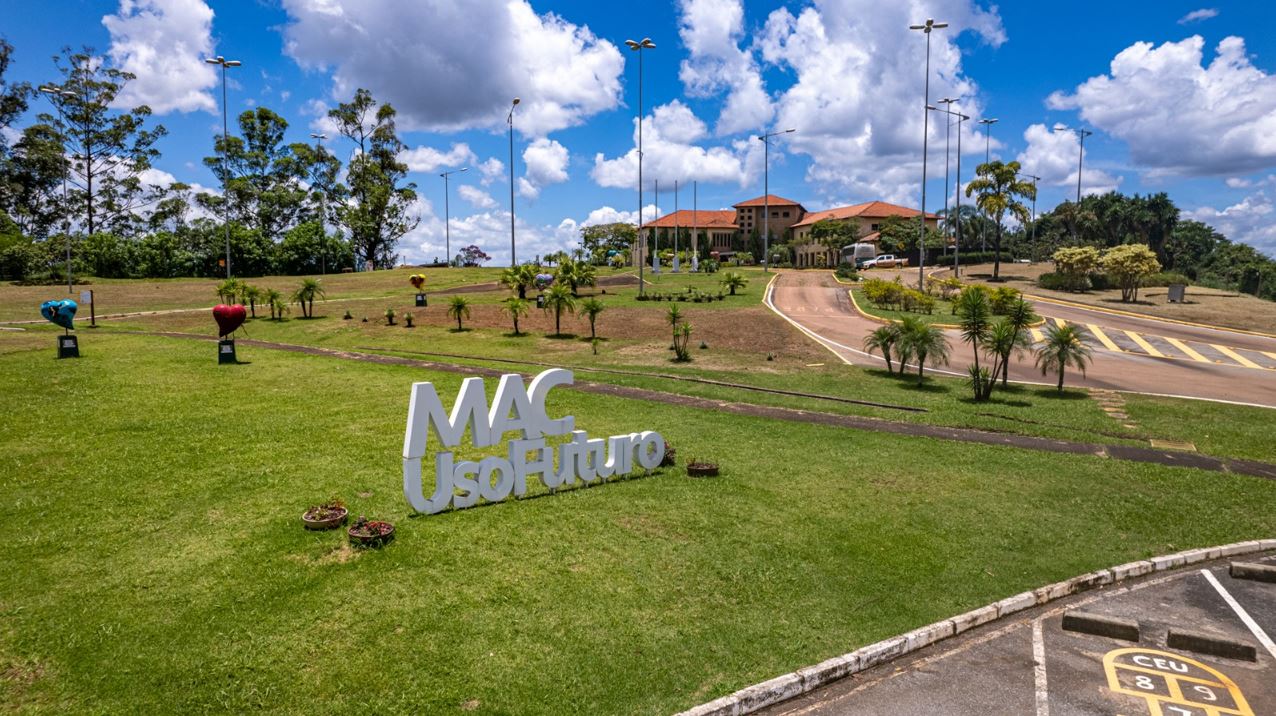 Entrada do complexo MAC Uso Futuro, com letreiro em destaque sobre gramado bem cuidado, esculturas artísticas coloridas, paisagismo e o prédio principal da antiga Mina de Águas Claras ao fundo, sob céu azul com nuvens.